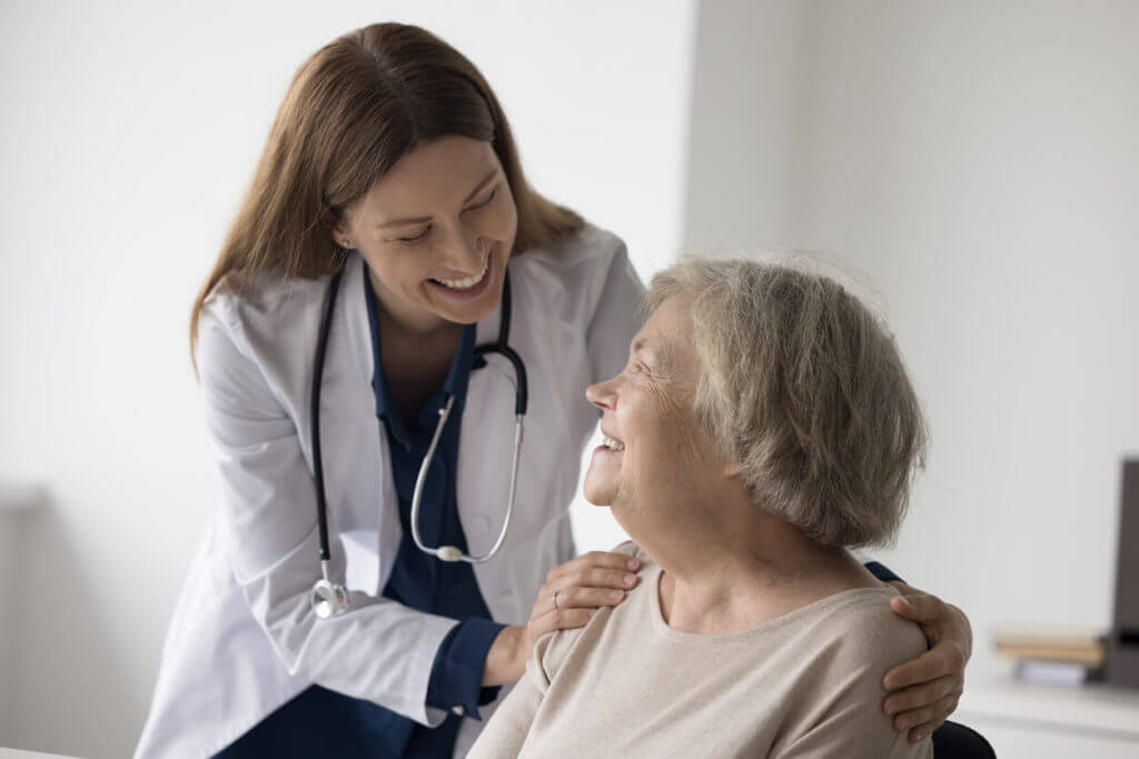 Cheerful female primary care physician wearing a stethoscope touches the shoulders of an elderly patient at the clinic.