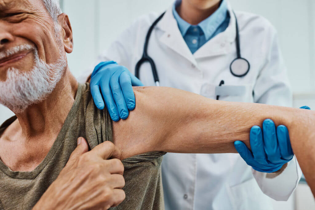 A senior man is experiencing pain from arthritis during a medical examination of his arm and shoulder at the doctor's office for arthritis treatment.