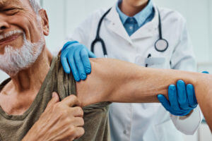 A senior man is experiencing pain from arthritis during a medical examination of his arm and shoulder at the doctor's office for arthritis treatment.